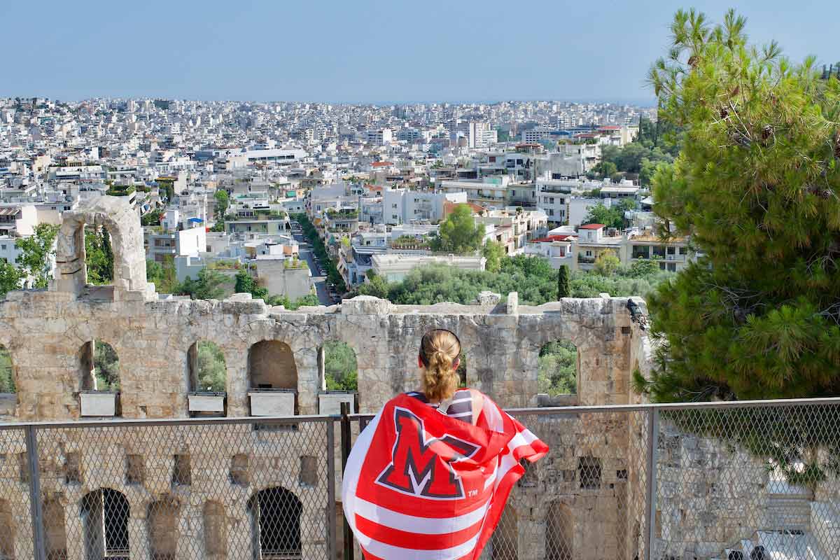 a student wrapped in a Miami University flag standing at a fence looking over Athens