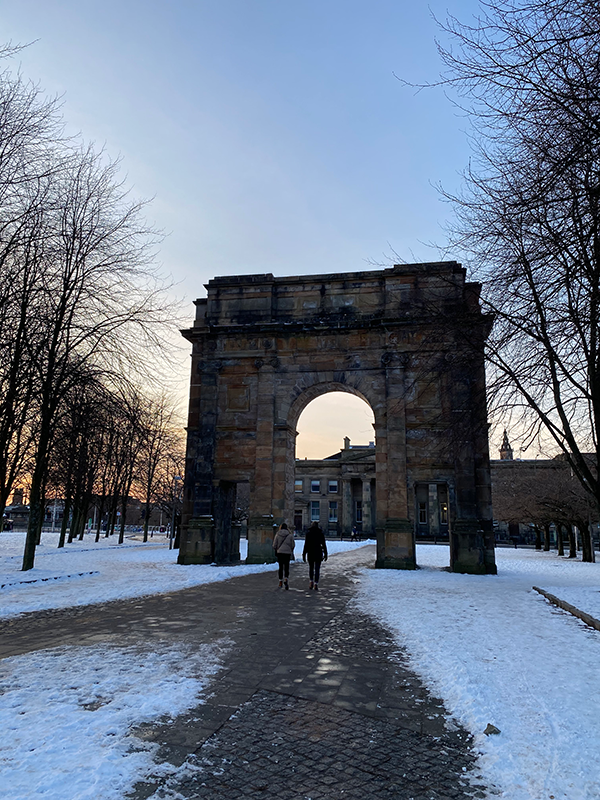 Two people walk beneath a large stone arch along a snow-covered path framed by leafless trees at sunset.