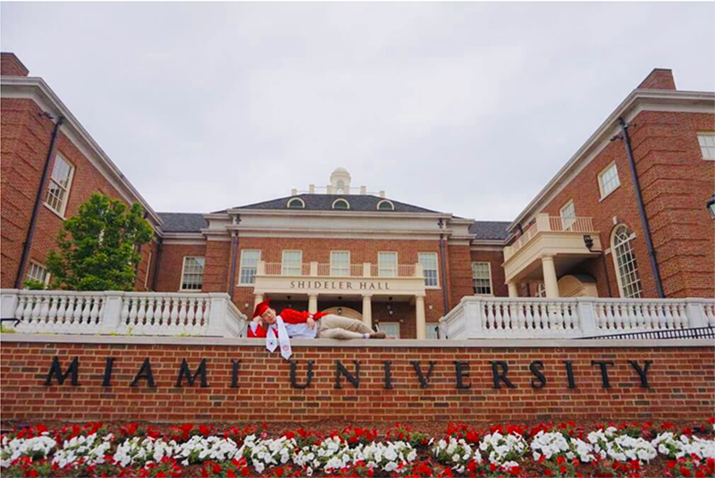James in cap and gown reclines in front of Shideler Hall