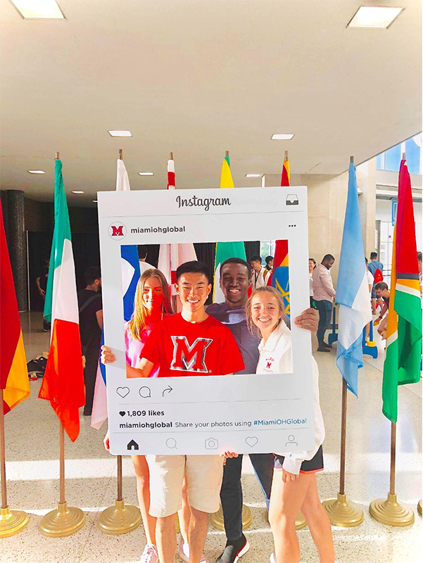 Regis and friends stand with an instagram frame in front of international flags