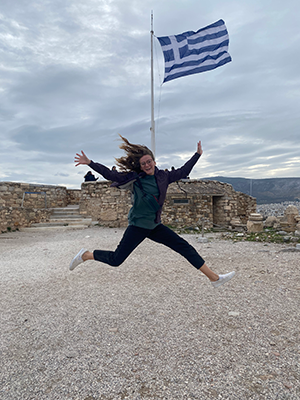 Rachel leaps in the air. The Greek flag is visible in the background