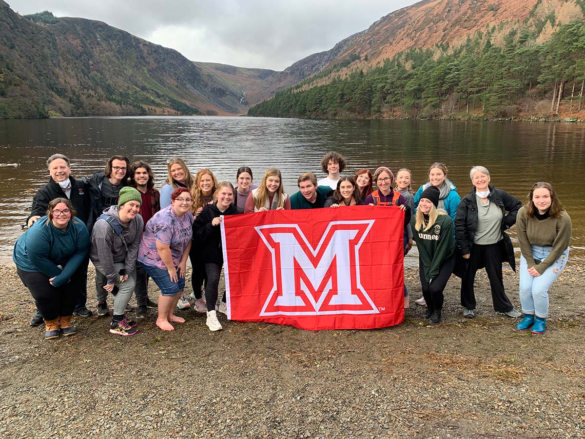 Students on a past Theatre in Dublin trip pose near a lake and hold up a Miami M flag