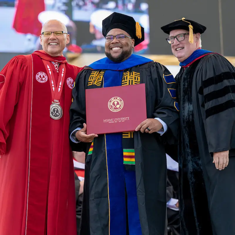 Graduate student with professor and Greg Crawford posing for photo holding diploma. 