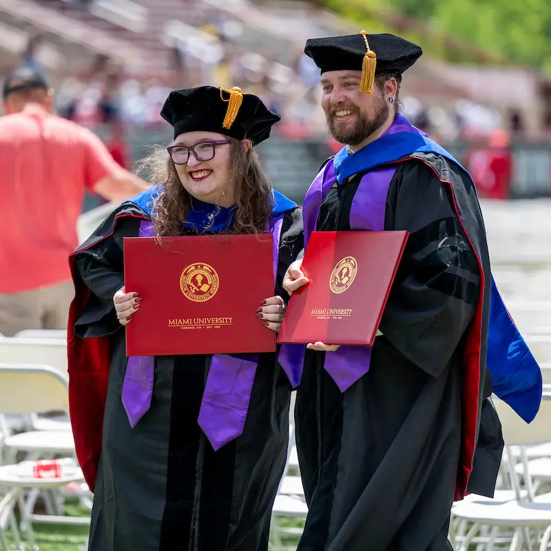 Two graduate students holding diploma outside, smiling, posing for photo. 