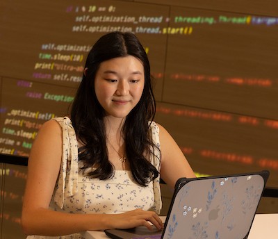Student working on laptop with coding language displayed behind her. 