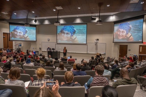 faculty and directors in front of lecture room with large screens displaying thesis presentation