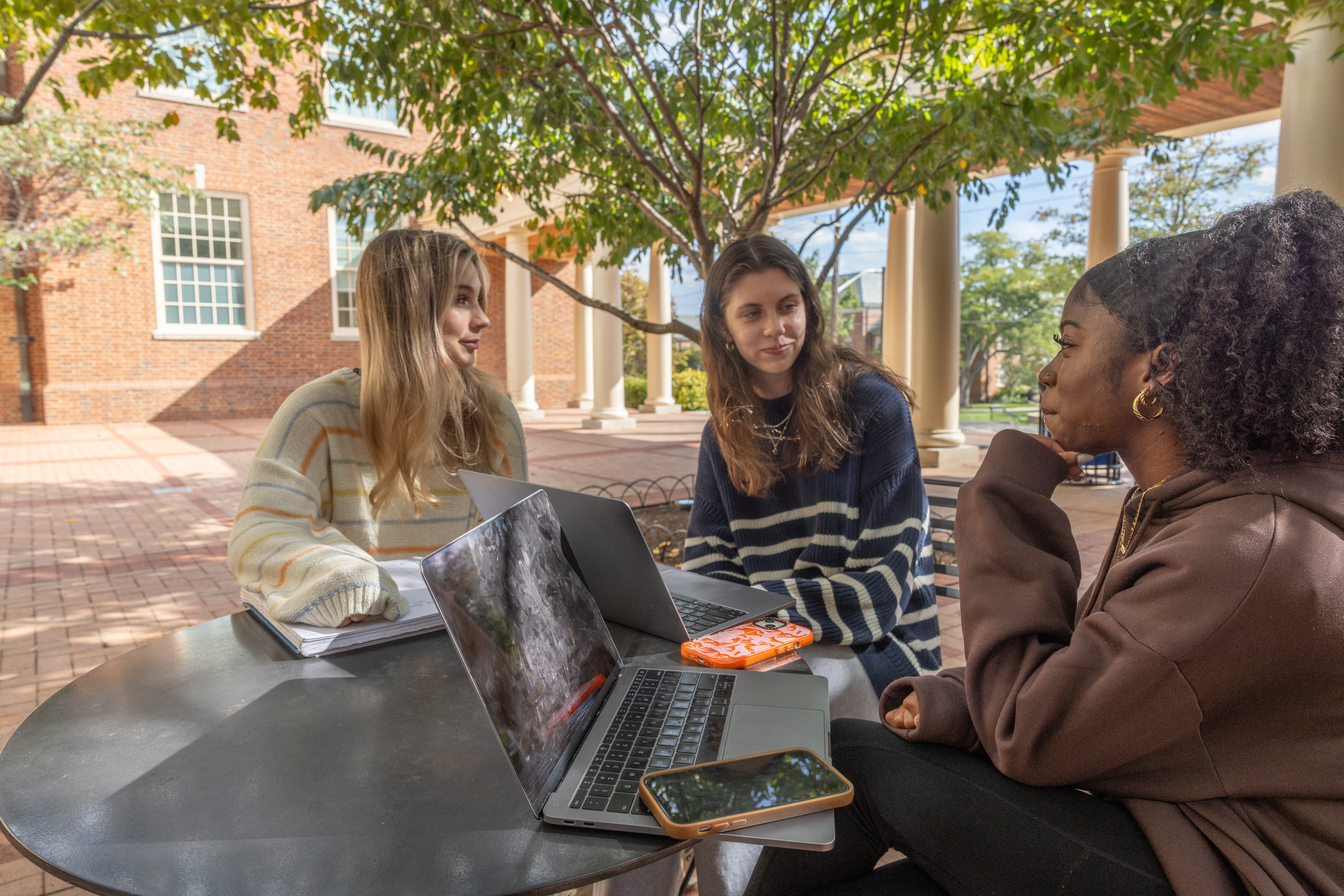 Three feminine presenting students sitting outside facing each other with their laptops open.
