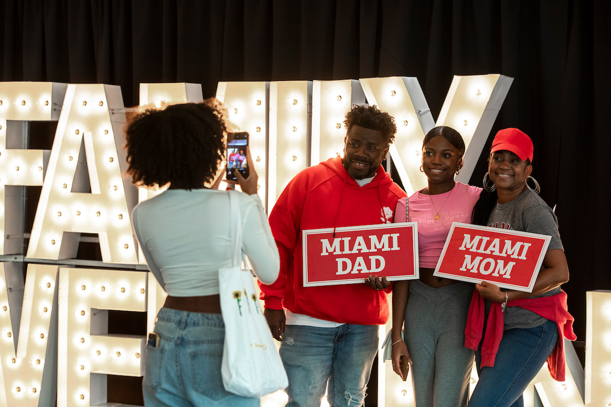 A family stands in front of a 'family weekend' banner, holding 'miami mom' and 'miami dad' signs