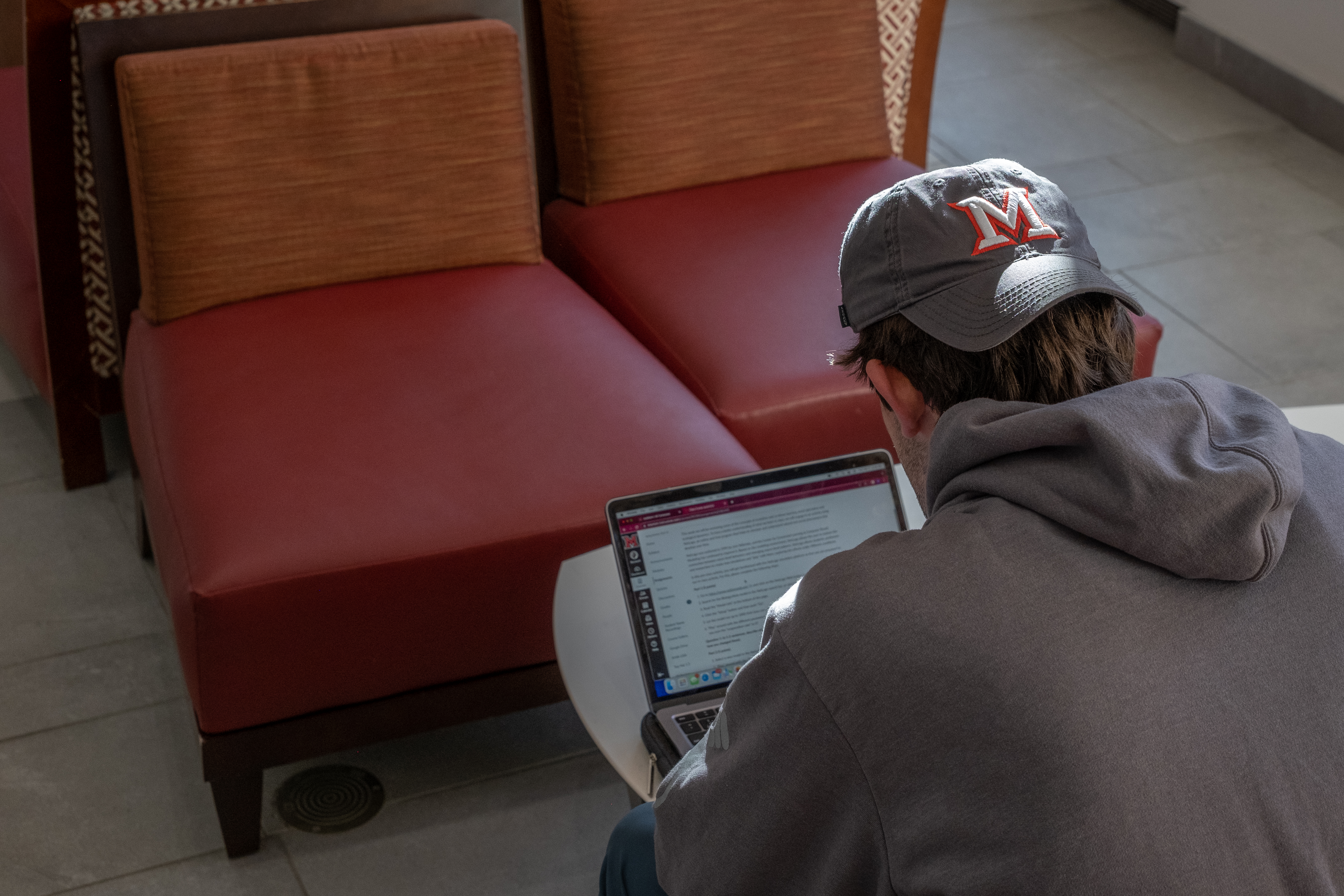 Shot of the back of a student facing away from the camera, sitting and regarding their laptop. The student wears a hat on backwards with the Miami symbol on it.