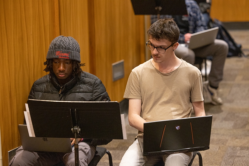 Two students sit on folding chairs on laptops. One student is looking at the other's screen.