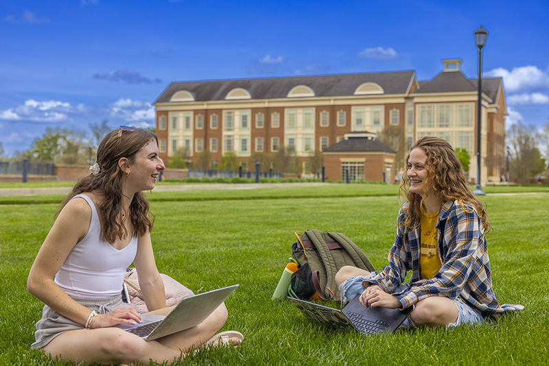 Two female-presenting people sitting on the lawn with laptops