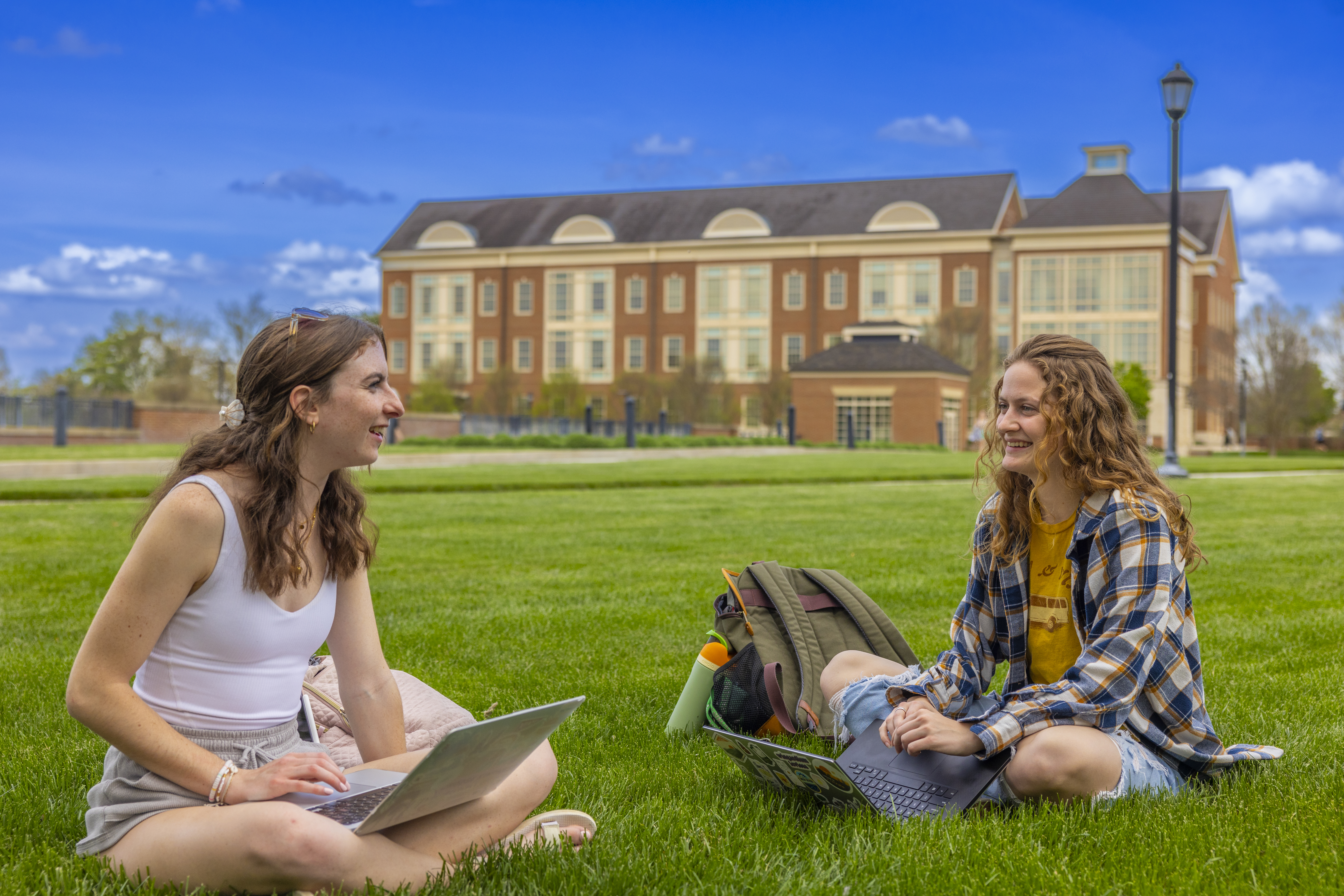 Tow feminine presenting students sitting outside on the grass with their laptops open in their laps.