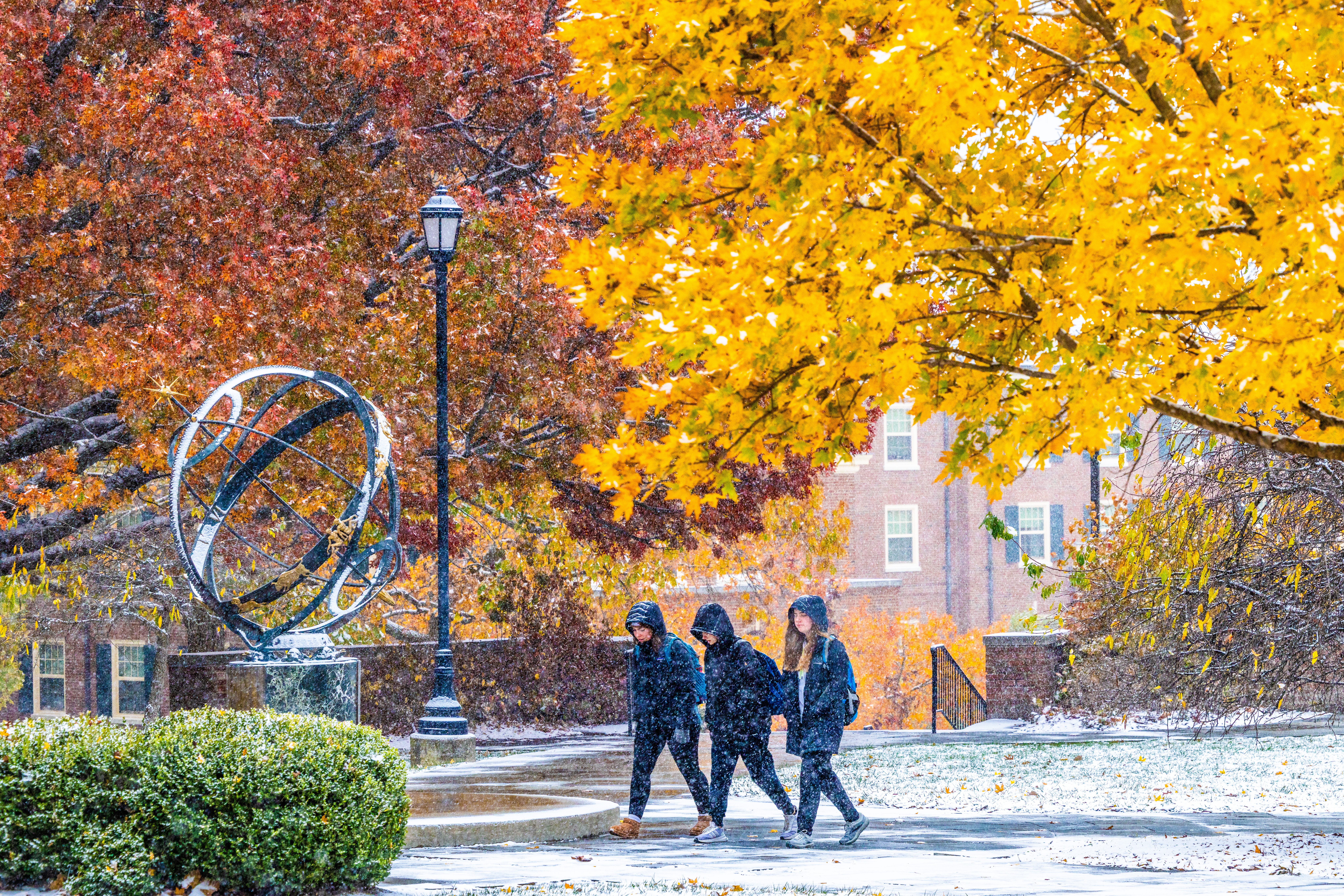 A group of students walking across campus past the Compass in the snow and orange leaves