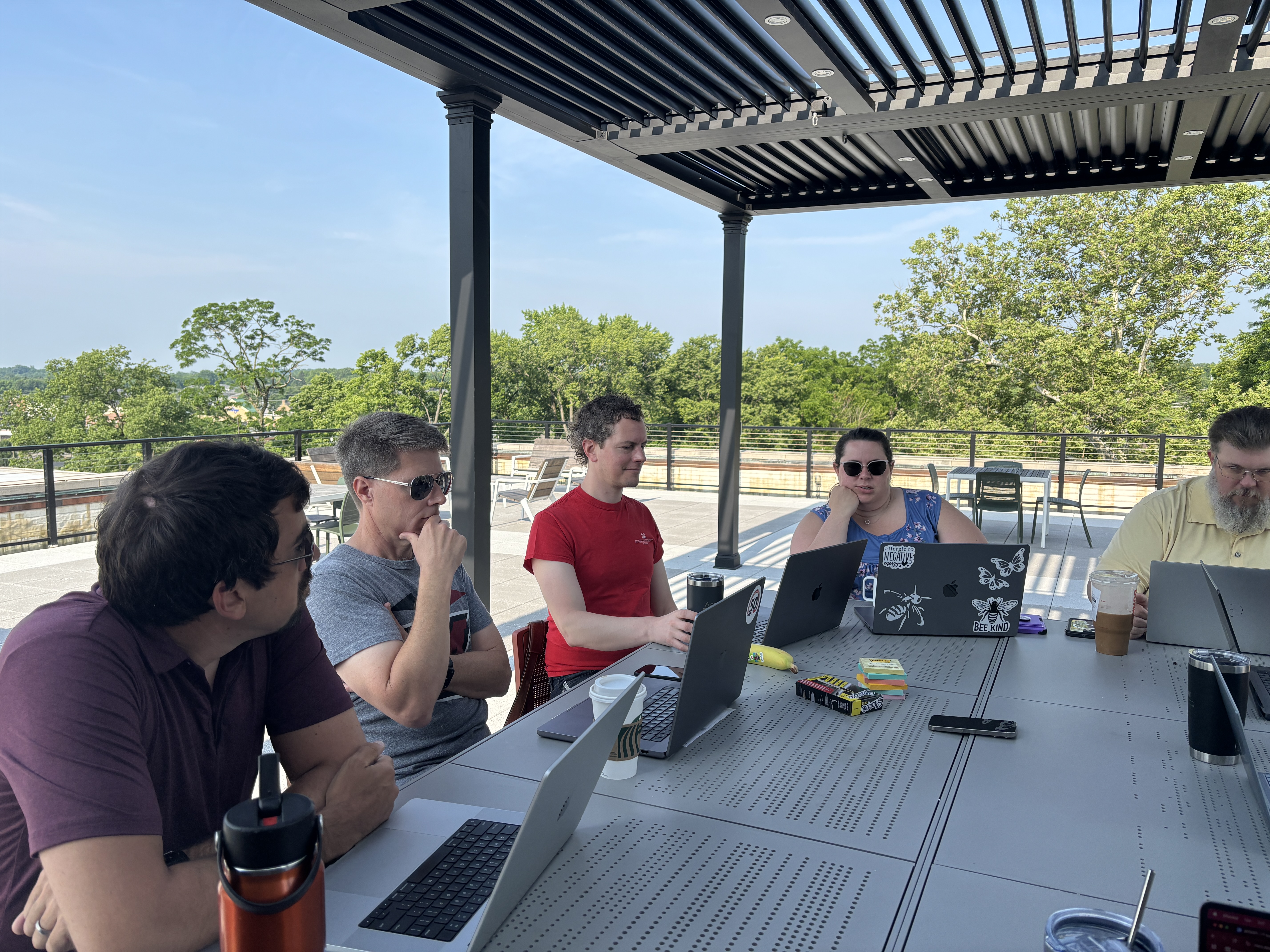 A group of people sitting around a table on their laptops outside. Some are wearing sunglasses. All appear deep in thought.