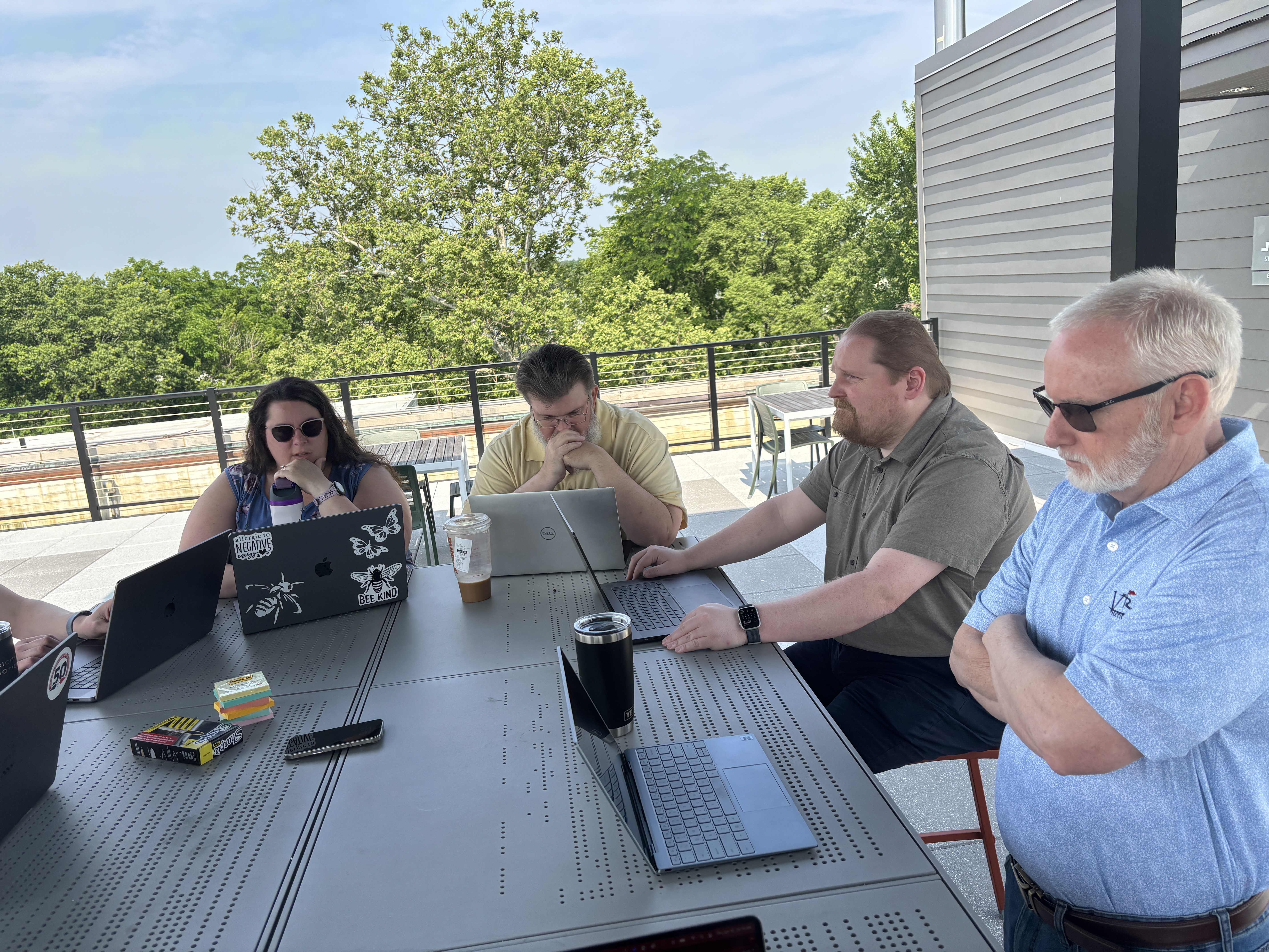 A group of folks sit around a table outside in the sunshine, some wearing sunglasses. They look at their laptops pensively.