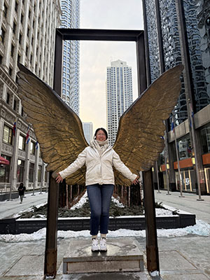 Ruowei standing in front of a large art display shaped like an angel's wings, framed in such a way that it looks like the wings are hers, on a city street.