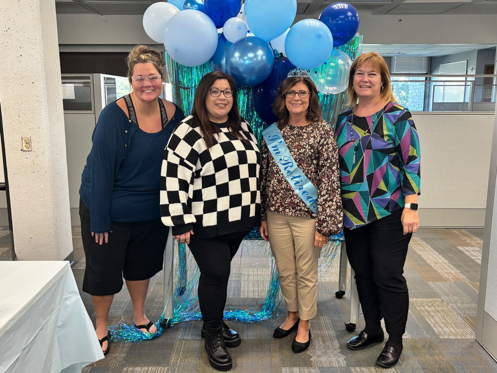 Kasey Arthur, Emma Lester, Sandy Keller, and Amy Moore stand in front of a blue background with balloons. Sandy is wearing a sash labeled 'I'm retired!'