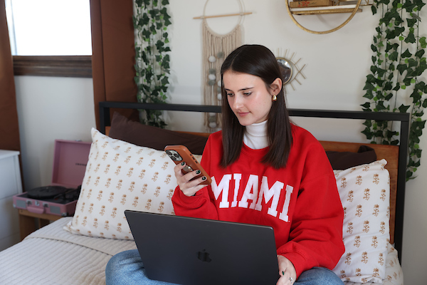 A student wearing a Miami sweater and using a laptop and smartphone