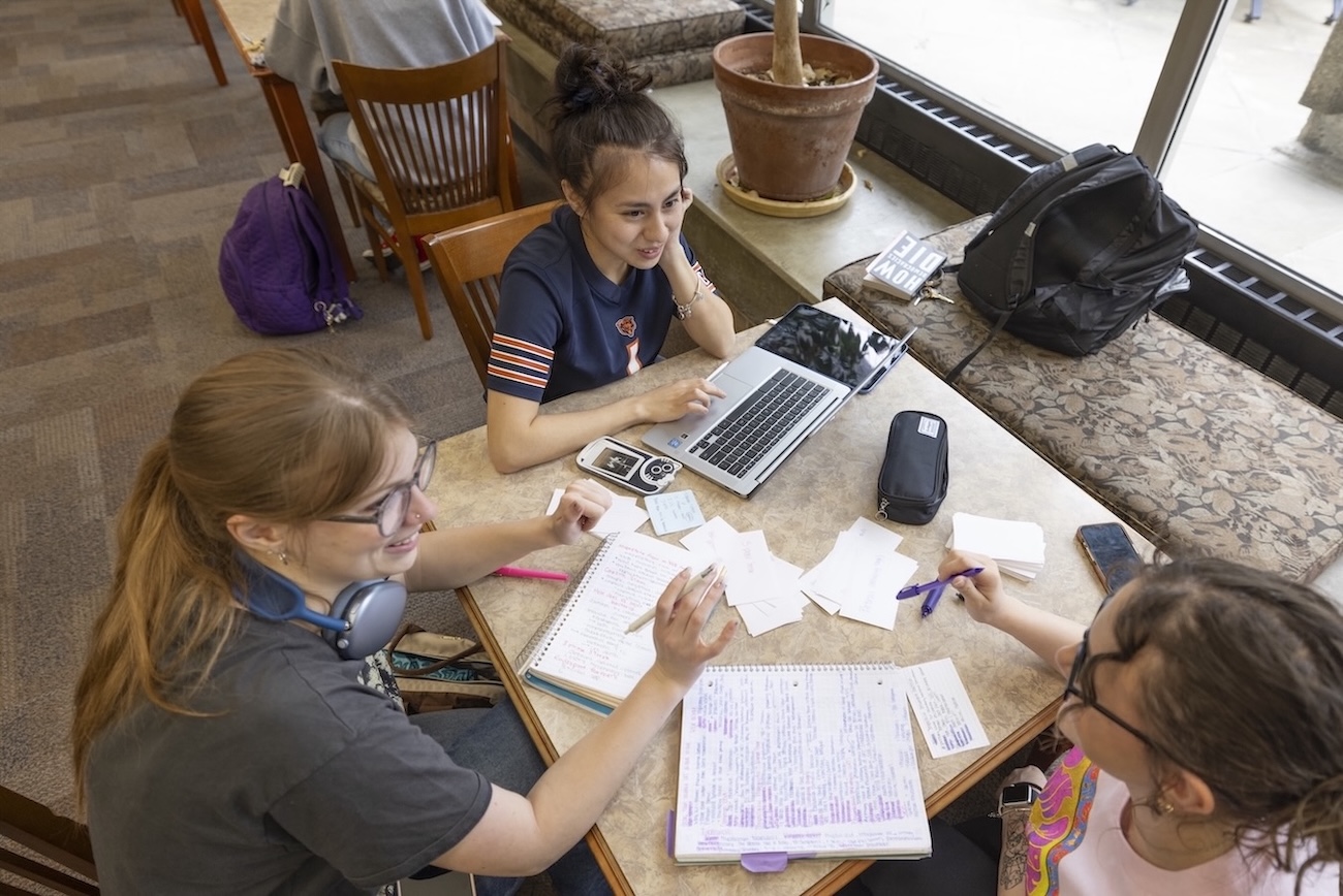 Three female students sitting around a table talking and taking notes. One student is using her laptop.