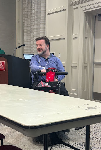 Tom Webb, a white man using a mobility chair, at the front of a classroom full of long white tables.