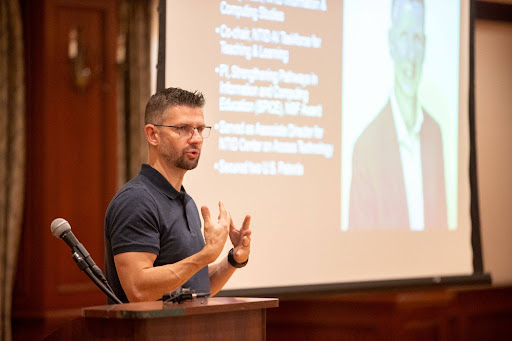 Brian Trager, a white man with short dark hair and glasses, stands at a podium in front of a projector showing slides, gesturing with his hands.