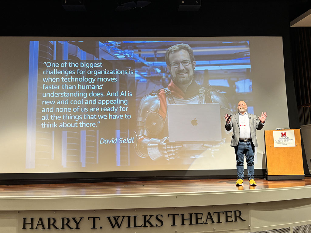 Brian Baute stands onstage in the Harry T Wilks auditorium in front of a slide that features a quote from David Seidl