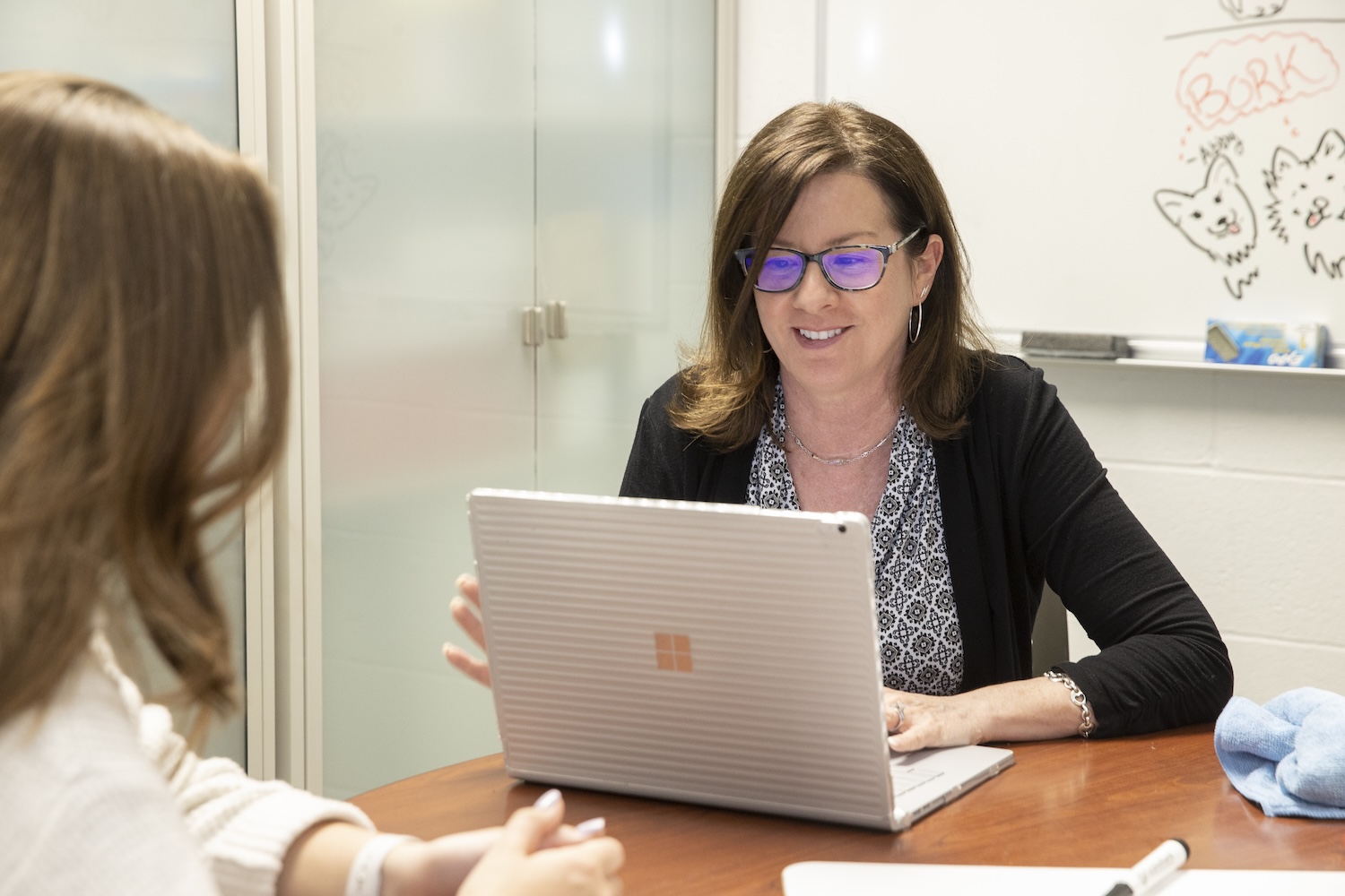 A female-presenting staff member wearing glasses sits at a table on her laptop. A whiteboard behind her has drawing of dogs that say BORK
