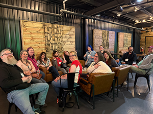 A group of IT professionals cheesing for the camera. Some sit on the couch and some in chairs. All are smiling and happy for each other's company.