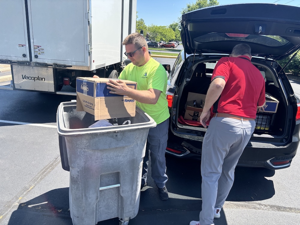 Two ShredFest volunteers unloading paper from the back of a car and loading it into a dumpster to get shredded.