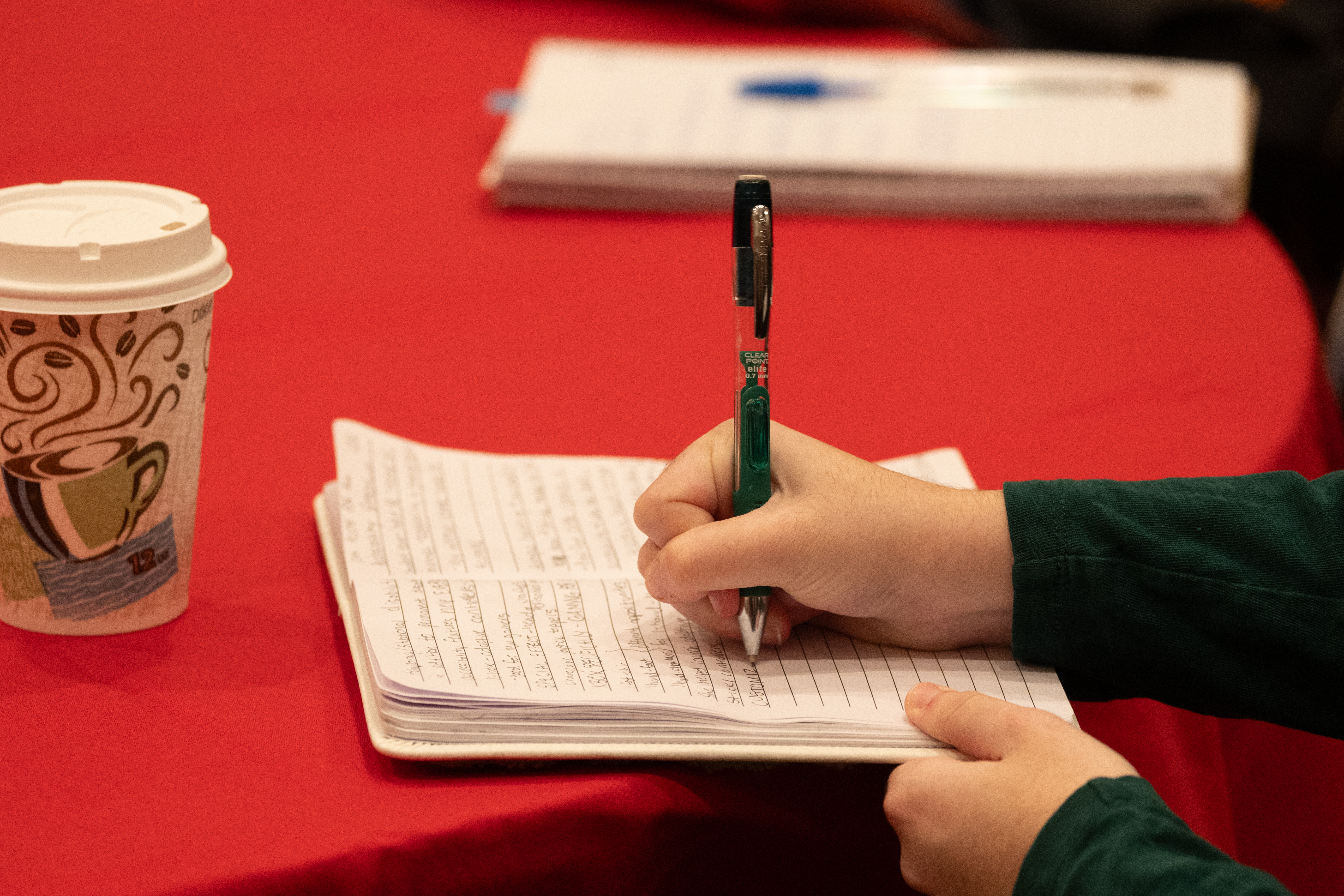 A person writing in a notebook. A cup of coffee sits on the table next to them.