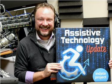 Josh Anderson wears headset and stands in podcast studio. He holds up a sign reading Assistive Technology Update