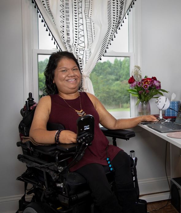 Liz Persaud is smiling and seated in her wheelchair next to a window.