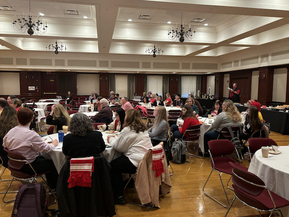 Attendees sit at round tables in the Heritage Room and listen to a presenter