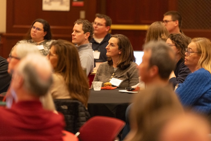 Attendees sit at round tables in the Heritage Room and listen to a presenter