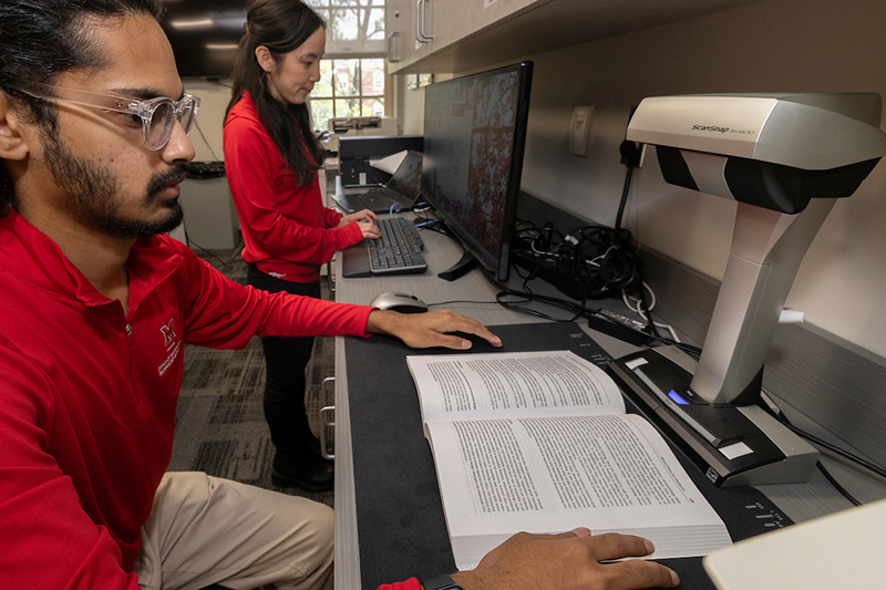 AccessMU student operates a book scanner. A staff member is seen in the background.
