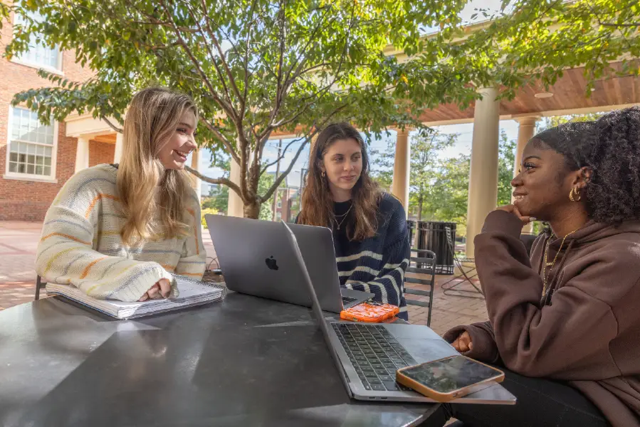 Several female-presenting young people sitting on a patio outside the armstrong student center. They are on laptops