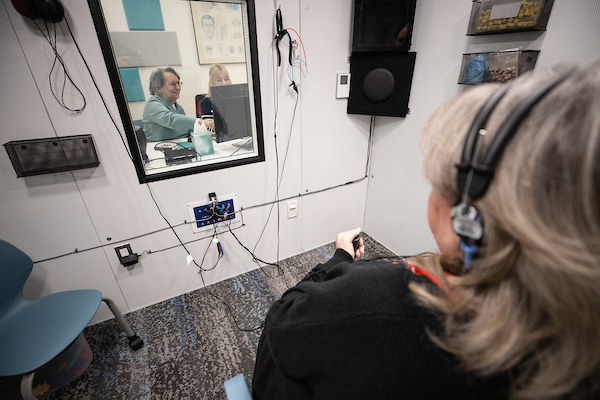 A woman wearing headphones is seated in a soundproof room, interacting with a person visible in a mirror across from her.