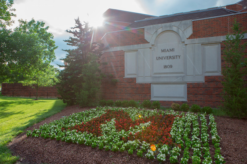 A brick wall reads Miami University. In front of the wall is the letter M made from flowers that are in bloom