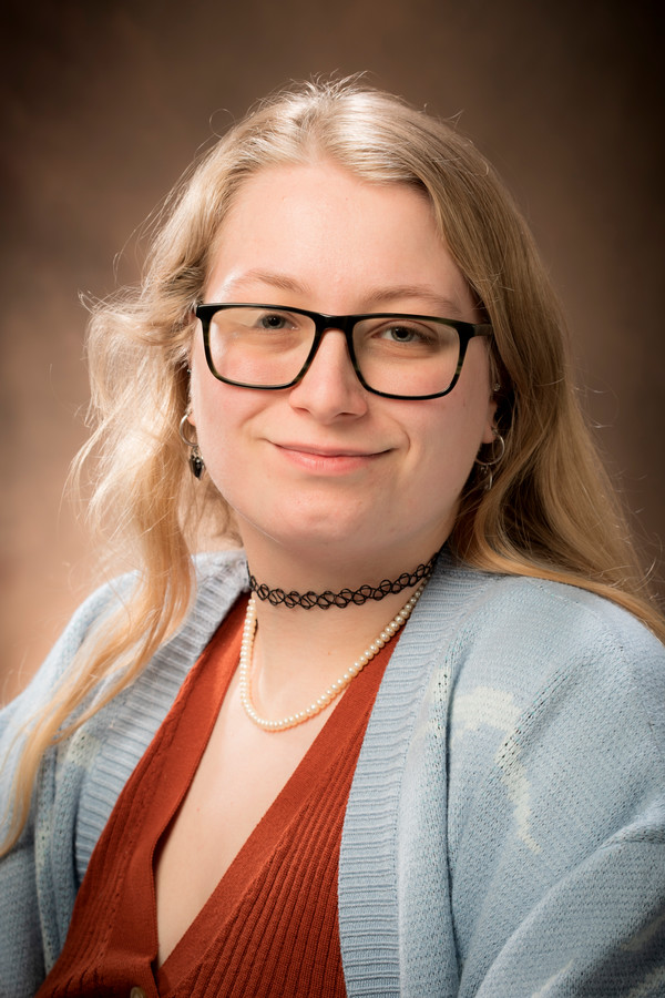 Person with long blonde hair, glasses, wearing a red shirt, and a blue cardigan.