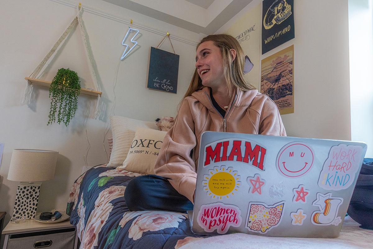 A smiling student sitting on their bed working on their laptop
