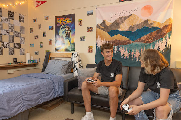 A male and female student sitting on a bed in a dorm room talking to one another