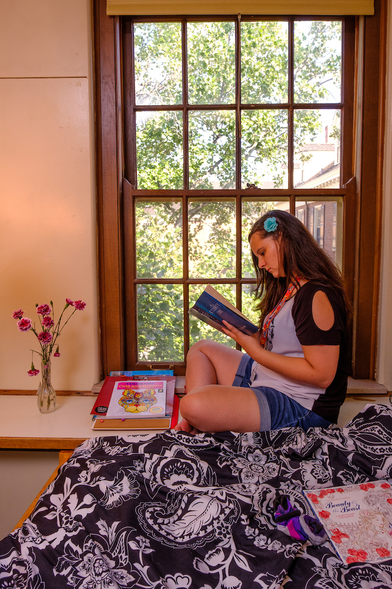 female student reading a book while sitting on a bed in a dorm room
