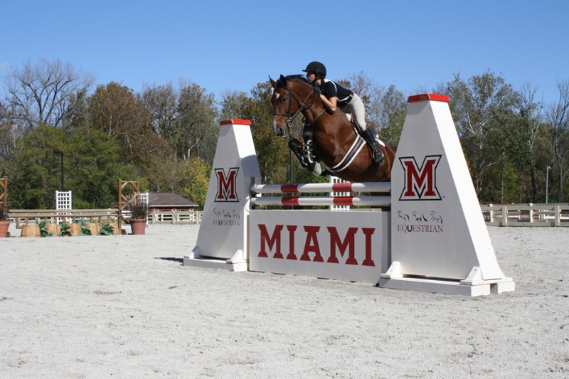 Student riding a horse that is jumping over a Miami branded hurdle.
