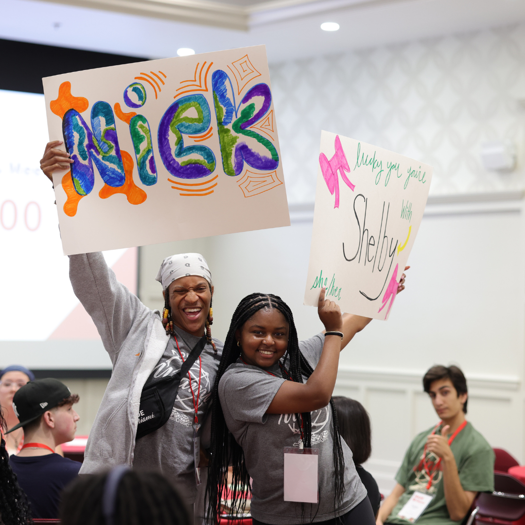 Two MADE Leaders holiding up poster board signs. One reads Nick in bubble letters and the other says Lucky you, you're with Shelby. 
