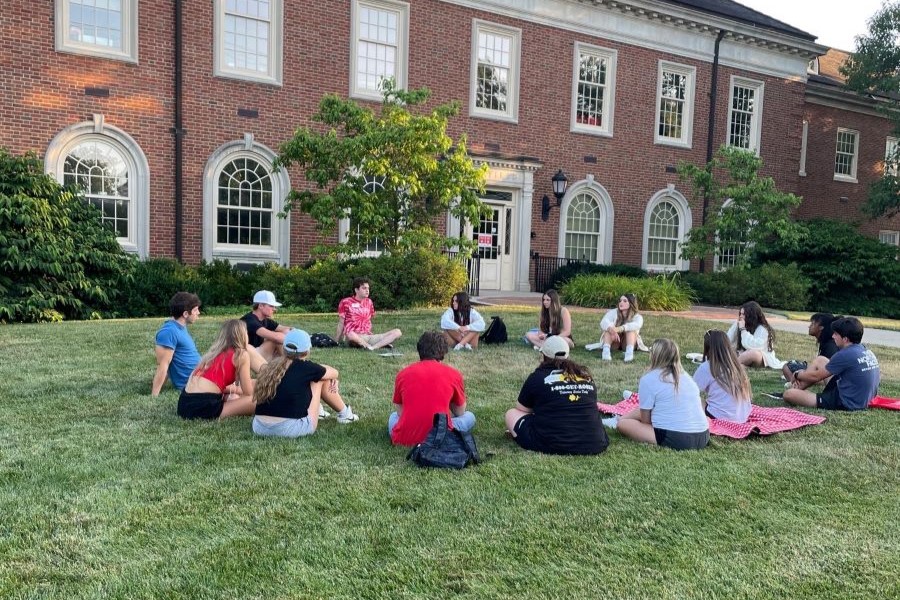 Group of about 15 students sitting in a large circle on the grass in front of Warfield Hall