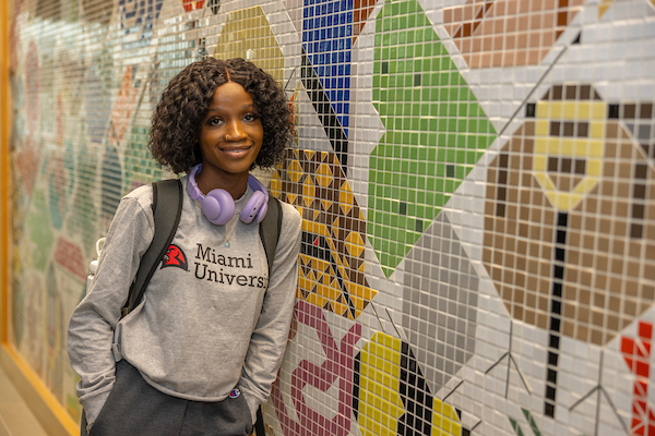 Student in Miami sweatshirt and backpack leaning against a tiled mosaic wall.