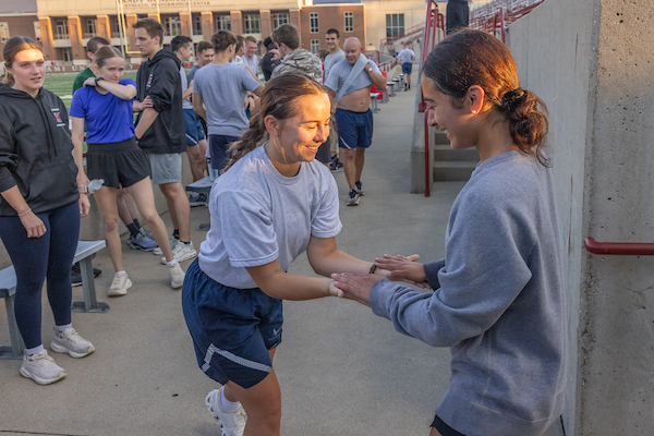 Two students facing each other with hands connected, one student palms face up and the other palms face down, with various other students in the background observing.