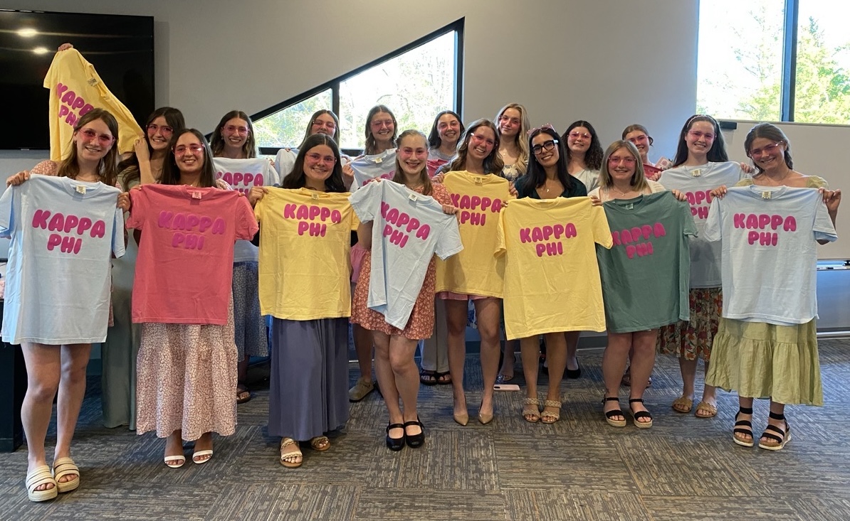 Kappa Phi sorority members are holding up colorful t-shirts that read "Kappa Phi."