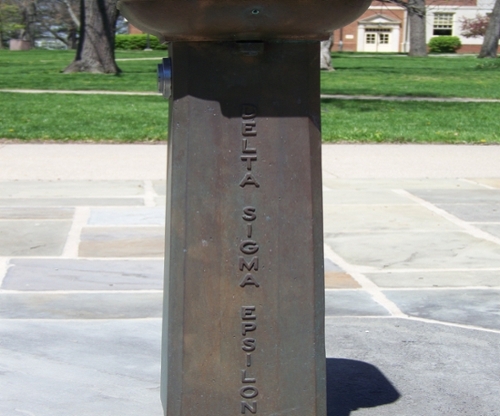 Metal base of a public water fountain that reads vertically Delta Sigma Epsilon.