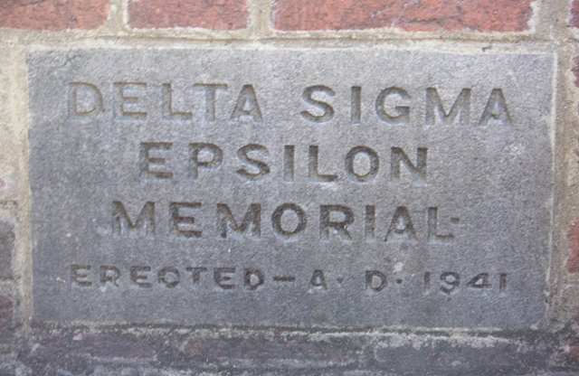 Metal base of a public water fountain that reads vertically Delta Sigma Epsilon.